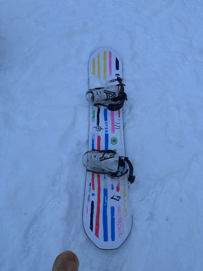 A snowboard lies in the snow. The board has a white background with colorful horizontal stripes and geometric designs. Bindings are attached near the center, appearing gray or white. The snow surrounding the board is textured. A tan shoe is slightly visible in the bottom right corner of the frame.