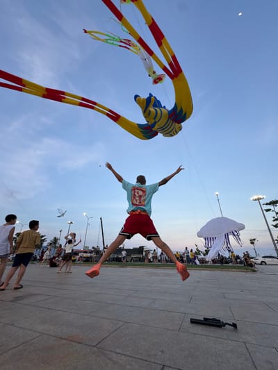 This image captures a lively outdoor event, likely a festival or gathering, with a focus on kite flying. In the foreground, a young man is captured mid-jump, his arms outstretched, wearing a light blue t-shirt with a red lobster graphic and red shorts. He's wearing bright orange sneakers. Behind him, a large, colorful kite resembling a dragon or bird with yellow, red, and blue elements dominates the left side of the frame, with long red and yellow streamers trailing. Further in the sky, a large white kite shaped like a jellyfish with purple accents floats serenely.
The setting appears to be a paved outdoor area, possibly a promenade or park, with a crowd of people visible in the background. The ground is made of large, light-colored rectangular tiles. In the distance, there are streetlights, some trees, and what looks like a stage or event setup with sound equipment. The sky is a clear, pale blue, indicating it is likely daytime, possibly late afternoon or early evening given the soft lighting. The atmosphere seems joyful and energetic, suggested by the dynamic pose of the young man and the presence of many spectators.
The location is identified as Quy Nhon Ward, Vietnam. There is no visible text in the image.