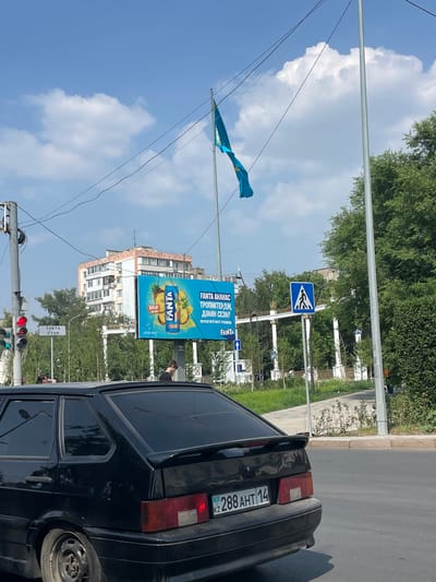 This media file captures a street scene in Pavlodar, Kazakhstan, under a bright, partly cloudy sky, suggesting a daytime setting. The foreground is dominated by the rear of a black sedan, likely a Lada, with a visible license plate reading "288 AHT 14 KZ". The car appears to be stopped, possibly at a traffic light, as indicated by a traffic signal visible in the lower left corner, showing red and green lights.
Behind the car, a large billboard advertising Fanta is prominently displayed. The billboard features an image of a Fanta can surrounded by pineapples and other tropical fruits, with text in both Kazakh and Russian. The Kazakh text reads "FANTA AHAHAC ТРОПИКТЕРДІҢ ДӘМІН СЕЗІНІ" which translates to "Fanta's new tropical flavor. Feel the taste." The Russian text below says "ПОЧУВСТВУЙ ВКУС ТРОПИКОВ", meaning "Feel the taste of the tropics."
Further back, a multi-story building stands with a light-colored facade, interspersed with trees. A pedestrian crossing sign is visible on a pole to the left of the billboard, indicating a crosswalk in the vicinity. A blue flag, possibly the flag of Kazakhstan due to its color and design, is seen fluttering on a pole to the left of the billboard and above the pedestrian crossing sign. The overall scene depicts an urban environment with typical street elements like traffic, advertising, and architecture.