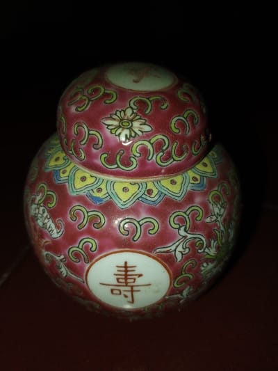The image displays a small, lidded ceramic jar, centrally positioned and illuminated against a dark, featureless background. The jar, predominantly deep reddish-pink, features intricate polychrome decorations. Its domed lid is adorned with green scrollwork and a prominent white flower with a yellow center. Below the lid, a band of yellow scalloped patterns with blue and red accents encircles the jar's upper body. The main body of the jar continues the reddish-pink base with green scrollwork motifs. On the front, a white oval cartouche contains a reddish-brown Chinese character, identified as '寿' (shòu), meaning longevity. Additional stylized white and green floral or scroll elements are visible on the sides of the jar. The object rests on a dark reddish-brown surface. No individuals, actions, or interactions are present. The precise urban location in Longmiang, Laos, cannot be determined from the visual evidence provided; the scene appears to be an isolated, indoor photographic setup.