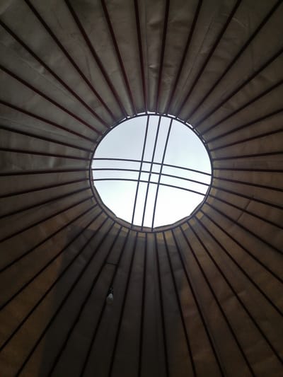 The image shows the interior ceiling of a yurt, likely located in or around the city of Taldyk, Kyrgyzstan. The ceiling is constructed with fabric stretched over a framework of wooden poles that radiate from a central circular opening at the top. This opening, a type of skylight known as a "tono" in Kyrgyz yurts, allows natural light to enter. The fabric appears to be a light-colored material, possibly canvas or felt, stretched taut between the poles. Some poles are visible as dark red vertical lines. A single light bulb hangs from a wire attached to one of the poles, suggesting the yurt is also equipped with artificial lighting. The overall impression is one of simple, functional construction, typical of traditional nomadic dwellings in Central Asia. The sky is visible through the central opening, indicating daylight. There are no people or animals visible in the frame.