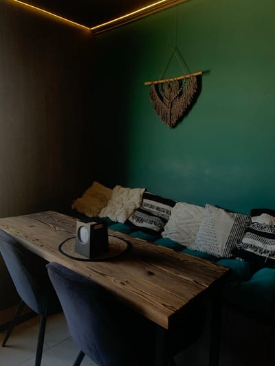 The image displays an unoccupied interior corner of a room in Yasinia, Ukraine. A long, rectangular wooden table with a distressed finish dominates the lower-center, flanked by two dark-upholstered chairs positioned to its left. Along the right, a dark teal-green wall features a matching upholstered bench seat with button tufting. Six decorative throw pillows are arranged on the bench: one textured mustard yellow, three textured cream/off-white, and two black-and-white patterned with fringe. Above the bench, a brown macrame wall hanging is suspended from a wooden dowel. A continuous strip of warm yellow LED lighting is affixed to the ceiling along the junction of the teal-green wall and the adjacent dark gray wall. Centered on the table, resting on a circular woven placemat, is a black, cube-shaped electronic device with a circular display and a visible "MOLEAP" label on its top surface. The floor is composed of light-colored tiles. No individuals are present in the scene.