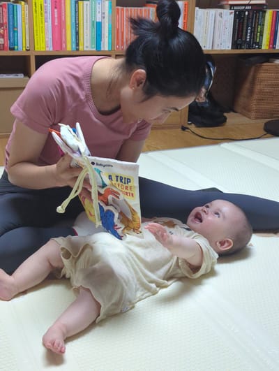 A woman with her dark hair in a bun sits cross-legged, facing a baby lying on a light yellow patterned surface. The woman holds a soft book open for the baby to view, the cover titled "A TRIP BEFORE BIRTH". The baby is fair-skinned and laughing, wearing a light-colored romper. The woman is wearing a light pink t-shirt. Behind the woman, a bookshelf is filled with books, including several titles by Richard Dawkins and one titled "Sapiens". Various other books are visible, including Montessori-related books and Chinese encyclopedias. A woven basket and a dark-colored bag are on the shelf above the books. The exact location in the city of Shunde District, China cannot be determined from the provided image.