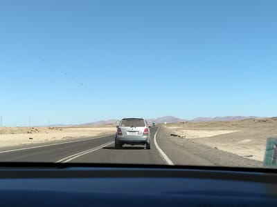 A silver Kia Sorento SUV, seen from behind, travels on a two-lane asphalt road in a desert landscape under a clear blue sky. The vehicle is covered in dust, indicating it has been driven in arid conditions. The road has white dividing lines, and a white line is also visible on the right shoulder. To the left of the road are sandy hills and sparse vegetation. In the distance, power lines and transmission towers are visible on the left horizon, and a range of arid mountains forms the backdrop. A blue truck is visible further down the road, ahead of the Kia. The scene appears to be along the Ruta 11-CH highway, a major route connecting Iquique to Oruro, Bolivia, passing through or near Pozo Almonte, Chile, known for its dry desert climate and mining operations. The image is taken from inside another vehicle, as indicated by the dark dashboard in the foreground.