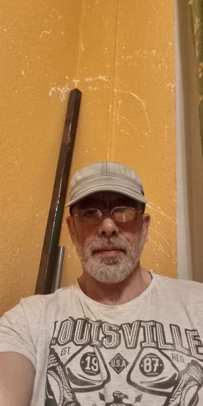 A middle-aged man wearing glasses and a light-colored baseball cap poses for a selfie against a textured, mustard-yellow wall. He has a gray beard and mustache and is wearing a light gray t-shirt with "LOUISVILLE EST. 19, U.S.A. 87 REG." printed on it in a black, stylized font. To the left of the man, a dark, metallic bar leans vertically against the wall. The lighting in the image is somewhat dim, casting soft shadows and highlighting the texture of the wall, which appears to have some white speckles and thin, white paint splatters. The scene suggests an indoor setting, possibly a workshop or garage, given the presence of the metallic bar and the rough texture of the wall. There are no indicators of the specific location within Wutha-Farnroda, Germany.