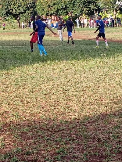 The image shows a grassy field with five young men playing a game of soccer at the Sokoine Stadium in Arusha, Tanzania. The field is covered in green grass, but it is patchy in places. There are some brown patches of dirt. The five players are wearing shorts and jerseys. The young men are wearing blue and red jerseys, all of them are running and kicking the soccer ball.  Two young men are wearing red shorts. One of the young men wearing red shorts is wearing blue socks.  The young men are interacting with each other. There are many people in the background watching the soccer match.  The stadium is surrounded by trees.  The weather is sunny.  The ground is dry.  The image appears to be taken from a distance.