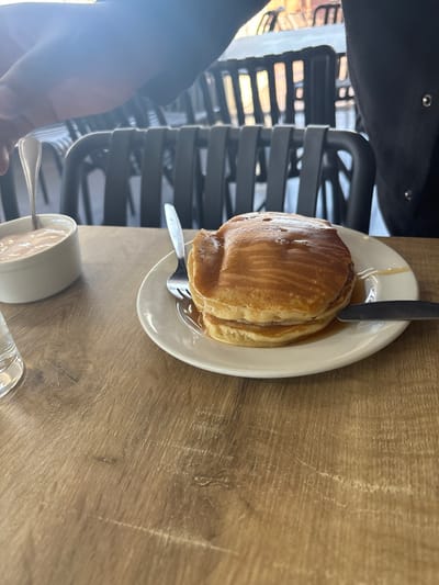 This image captures a meal setting in a dining establishment located in Port Alfred, South Africa. The primary subject is a stack of three golden-brown pancakes, heavily drizzled with a clear, golden syrup, presented on a white circular plate centrally positioned on a light brown wooden table. A silver fork is positioned to the left of the pancake stack, and a silver butter knife is to the right. To the left of the plate, a small white oval ceramic dish holds a light pink or orange creamy condiment, with a silver spoon resting inside. A partial clear glass is visible on the far left. Two individuals are partially visible: a person on the left with a dark-skinned forearm and hand reaching towards the condiment dish, and a person on the right, showing a dark-clothed shoulder and arm with a visible snap fastener. The background reveals multiple dark-colored chairs with vertical slatted backs, arranged in rows, indicating a restaurant or cafe environment. Bright light from an indeterminate source illuminates the scene from behind the foreground subjects.