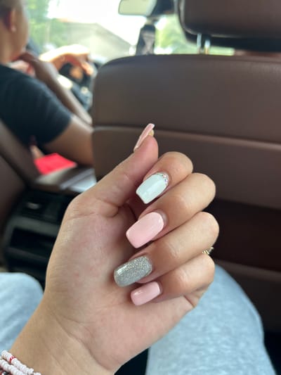 The image presents a close-up view of a person's right hand, showcasing a fresh manicure. The nails are a medium length with a square or squoval shape. The color scheme is varied, featuring pale pink on the thumb and pinky finger, a light blue or mint green on the index finger, and a sparkling silver glitter on the middle finger. The light blue nail is adorned with a line of small, clear rhinestones or gems placed along the cuticle. A gold-toned ring is visible on one of the fingers. On the wrist, a multi-strand beaded bracelet, primarily with white/pink and some red beads, is worn.

The background is out of focus, suggesting a shallow depth of field. It appears to be the interior of a vehicle, likely a car, with soft brown upholstery visible. Bright daylight streams in from the left side of the frame, illuminating blurry green foliage visible through a window. Partially visible in the background, out of focus, are shapes that suggest another person or parts of the vehicle's interior. The setting indicates that the photo was taken during the daytime, likely on a sunny day given the bright light. There is no visible text in the image.