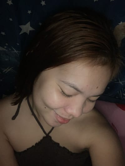 A close-up, overhead shot shows a person lying down on a blue blanket with white stars. The person's head is tilted down and to the right, with their eyes closed. Their brown hair is slightly damp and falls around their face. They are wearing a dark, ruffled tank top. The lighting is dim, casting shadows on their face and body. The image does not provide any information about the location within the city of Kiblawan, Philippines.