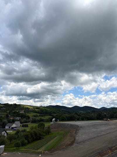 The image shows a construction site with a large, cleared area of dark gravel. In the background, there are rolling green hills dotted with houses and trees. The sky is filled with dramatic, dark clouds, with patches of blue sky and white, fluffy clouds peeking through. A yellow construction barrier is visible along the edge of the cleared area. Further in the background, a yellow construction vehicle, possibly a dump truck, is parked. The landscape suggests a rural or semi-rural setting, possibly in Stróża, Poland, given the context. The overall impression is one of an overcast day with a construction project underway in a scenic, hilly environment.