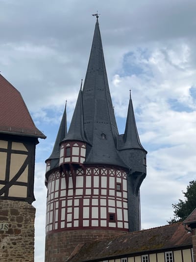The image depicts a close-up, vertically oriented view of a historic building with a prominent, pointed spire, characteristic of medieval architecture. The spire is covered in dark gray slate tiles, and it tapers to a sharp point. Below the spire, the building features a section with a distinctive half-timbered design, characterized by white infill panels and dark brown wooden beams arranged in a grid pattern. Some of these beams form decorative "X" shapes. Small, dark windows are visible within the white panels.

The building's construction also includes red brickwork. Adjacent to this structure, another building with a red tiled roof and a stone facade is partially visible. The sky above is filled with scattered white clouds against a pale blue background, suggesting it is daytime with variable weather.

The scene appears to be a tourist attraction or a historical site, possibly a castle or a well-preserved old town area, given the architectural style. There is no visible activity or people in the frame. The lighting suggests it is likely mid-day. No text is clearly legible in the image. The location context provided is Neustadt, Germany, which aligns with the style of architecture often found in that region.