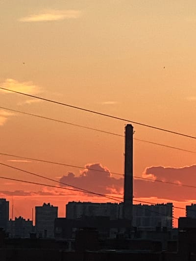 The image depicts a city skyline at dusk or dawn, with a predominant orange and yellow hue across the sky. In the foreground, silhouetted buildings form a dark band across the bottom of the frame. Above these, a tall, slender industrial smokestack stands prominently in the center of the image. Several power lines crisscross the sky. Fluffy clouds, tinged with the warm colors of the sunset or sunrise, are visible behind the smokestack and the buildings. The scene appears to be an urban or industrial area, possibly near Ladozhskoe Ozero, Russia, given the context. There are no people or discernible actions taking place in the image.