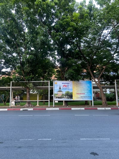 In Tha Khlong Town Municipality, Thailand, a billboard is displayed in front of lush green trees and a street. The billboard features Thai text and images of a university building. Two individuals, a woman and a child, are walking past the billboard. The scene is captured from a street-level perspective, with the road and sidewalk in the foreground. The environment appears to be an outdoor public space, possibly near a university campus.