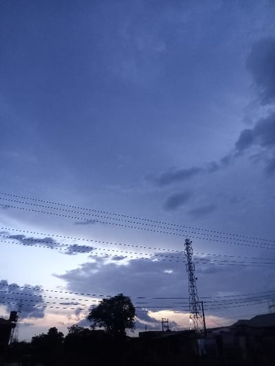 The image shows a view of the sky from a residential area in Ita Oniyan, Nigeria. It's an evening shot with a blue sky and a few wispy clouds. In the foreground, there are multiple power lines running horizontally across the sky, converging towards a cell tower. The tower stands tall against the setting sun, silhouetted against the fading light. A few trees are visible, also in silhouette, along the bottom edge of the frame. The scene is peaceful and serene, suggesting a quiet moment in a tranquil setting. There are no people visible in the image.