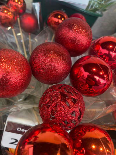 This image depicts a collection of red spherical ornaments, likely intended for festive decoration, displayed in a retail or market setting within the city of Aiguilhe, France. The foreground and midground are dominated by these ornaments, showcasing variations in texture and finish. Several large ornaments are covered in fine red glitter, presenting a matte yet sparkling appearance. Adjacent to these are multiple smooth, highly reflective red spheres that mirror ambient light and indistinct shapes. One prominent ornament exhibits an openwork or filigree design, also finished with red glitter. In the background, some smaller red spheres are visible attached to thin wooden sticks and individually encased in clear plastic wrapping. A dark green container or basket is partially visible towards the upper right, and a white tag with partially obscured text, including the letters "Dé" and the number "2," is visible in the lower-left background, also partially covered by a plastic wrap. No individuals are present in the scene.