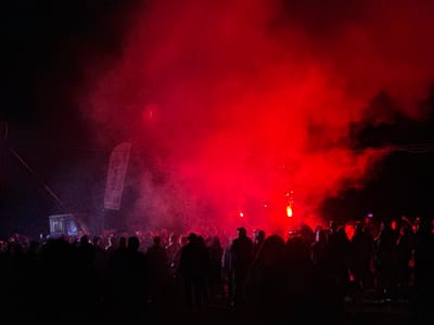 The image captures a nighttime outdoor gathering in Большая Кивара, Russia. A dense crowd of individuals occupies the lower and mid-ground, predominantly appearing as dark silhouettes against a backdrop of intense red light and pervasive smoke or haze. The red illumination emanates primarily from the right side of the scene, where multiple individuals within the crowd are observed holding and emitting bright red flares or pyrotechnic devices, which contribute to both the light and the smoke. In the upper-left quadrant, a dimly lit, box-shaped vehicle, possibly a utility van or small truck, is present. Above this vehicle, a vertical white banner is visible, displaying partially discernible text, which appears to be Cyrillic. Multiple dark wires or cables stretch across the upper-left portion of the frame. The ground beneath the crowd is indistinct but appears unpaved. The scene portrays a mass assembly under specific lighting conditions produced by the crowd itself.