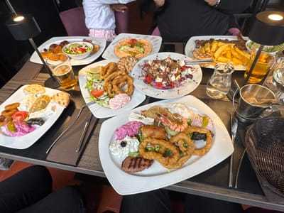 The image depicts an indoor dining scene at a restaurant, identified as being located in Evergem, Belgium. A wooden table with dark gray placemats is laden with numerous dishes, indicating a communal meal for multiple individuals.

At least three individuals are partially visible. In the immediate foreground, the legs and lap of a person wearing black trousers are visible. To the left, a child's torso in a white top and pink patterned trousers is partially obscured. To the right, an adult's arm and torso in a black top are seen.

The table is densely populated with a variety of Mediterranean-style dishes served on white plates:
*   **Foreground Right Plate:** Features grilled shrimp, fried calamari rings, a grilled meat patty, a small pastry roll, and various dips (pink beetroot-like, white tzatziki-like, and crumbled feta cheese) served on a bed of lettuce with an olive and a green pickled pepper.
*   **Mid-Left Plate:** Contains a substantial portion of fried calamari rings, a side salad with lettuce, cucumber, tomato, and a lemon wedge, accompanied by a pink dip and pita bread triangles.
*   **Mid-Right Plate:** A Greek salad with large chunks of feta cheese, red onion, tomatoes, olives, red bell pepper, and a generous sprinkle of dried herbs/spices.
*   **Background Left Plate:** Displays grilled meat pieces or skewers, roasted potato wedges, and a small side salad in a bowl, with pita bread.
*   **Background Middle Plate:** A serving of hummus or a similar spread, garnished with cucumber slices, tomato, and a green olive/pepper, surrounded by pita bread.
*   **Background Right Plate (partially visible):** Appears to contain grilled meat and a significant portion of french fries, alongside a small green salad.
*   **Foreground Left Plate (partially visible):** Shows fried cheese (saganaki style) and pita bread.

Beverages include at least two glasses of golden-colored beer, a small glass carafe of water, and a silver ice bucket with tongs. Two black, modern, cylindrical table lamps with illuminated tops provide focused lighting on the table. Cutlery (forks and knives) is neatly arranged on the placemats. A dark woven basket, likely for bread, is visible on the far right. The floor in the lower left corner shows reddish-brown tiles. The background is dimly lit and features purple upholstered chairs.