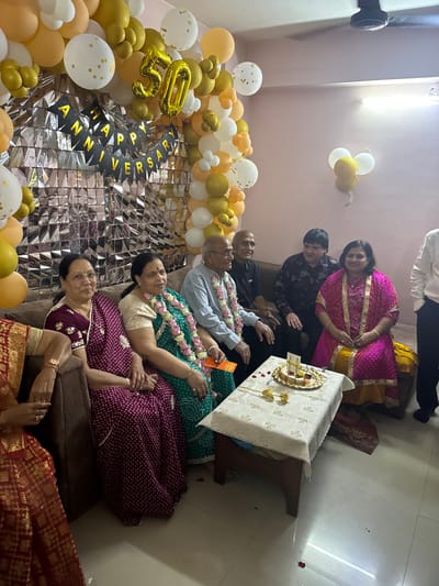 The scene depicts an indoor celebratory gathering in a residence located in Bidhannagar, India. The background is extensively decorated with a silver holographic tinsel curtain, a black banner bearing "HAPPY ANNIVERSARY" in gold lettering, and an elaborate arch of gold, white, and orange balloons, prominently featuring a large gold foil "50." Seven individuals are visible, with one partially obscured on the far left. On a brown upholstered sofa, two older women and an older man are seated; the man and the central woman wear floral garlands around their necks, and the woman holds an orange envelope. Another older man and a younger man are seated on a dark brown sofa behind a central table. To their right, a woman in a pink and yellow garment is seated on the floor, and a person in a white shirt is partially visible standing on the far right. A small rectangular wooden table, covered with a white patterned tablecloth, occupies the foreground. Upon the table, a silver tray holds multiple small dessert items, flanked by two golden decorative pieces and scattered red rose petals. The floor is white-tiled, also with scattered red rose petals. A ceiling fan is visible in the upper right. The individuals are generally facing forward, appearing composed for a photograph amidst the festive decor.