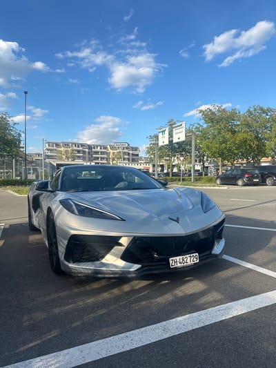 The image displays a silver or light gray Chevrolet Corvette C8 parked on asphalt, likely in a parking lot or by a roadside, under a bright blue sky with scattered white clouds. The car is the primary subject, viewed from its front-left side, showcasing its sleek design and headlights.
Visible on the front of the car is a Swiss license plate, "ZH-482 729," which includes the text "CORVETTECENTER.CH" on its holder, indicating registration in the canton of Zurich. Strong sunlight casts distinct shadows on the asphalt, suggesting it is daytime.
In the background, to the left of the vehicle, multiple road signs are visible. One sign points right to "Schwerzenbach" and "Dübendorf," while another, below it, indicates "Uster" to the right. Further behind, residential or commercial buildings with several stories are present, interspersed with green trees. Another sign, partially visible, reads "Ausfahrt" (German for 'Exit'). The overall setting confirms the location context of Volketswil, Switzerland, as indicated by the place names on the signs and the Zurich license plate. No people are visible, and the car appears to be stationary.