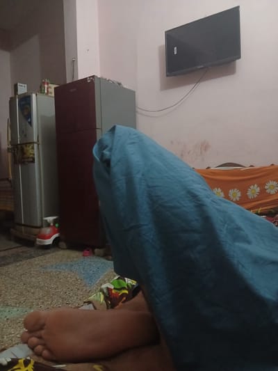 This is an indoor, eye-level, medium shot photograph taken in what appears to be a bedroom. The scene is captured in a low-light environment, with a dominant presence of a blue sheet or blanket covering a significant portion of the frame from the bottom right upwards and to the left. A bare human foot, with visible toes and sole, is positioned in the lower left corner, partially covered by patterned fabric.
In the background, to the left, are two refrigerators, one silver and one dark red, standing side-by-side. A small red and white toy car is placed on the floor in front of the refrigerators. Further to the left, a portion of a lighter-colored appliance, possibly another refrigerator or a cabinet, is visible. Above the refrigerators, a dark rectangular object, likely a television, is mounted on a light pink wall. The wall has some red marks near the top right. The floor appears to be tiled with a speckled pattern of brown and blue. An orange blanket with white daisy patterns is spread on a surface behind the blue sheet. The overall impression is a domestic setting within a residential space in Peshawar City Tehsil, Pakistan.