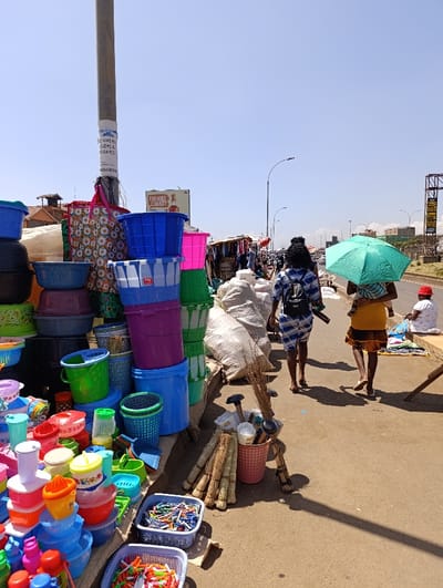 This image depicts a bustling street market scene in Githurai, Kenya, under a bright, clear midday sky, indicated by the sharp shadows cast on the ground.
The foreground is filled with an extensive display of colorful plastic household items, including numerous basins, buckets, food containers, storage bins, strainers, and smaller items like toys or clothes clips. These goods are stacked high on tables and arranged in baskets. Alongside these, bundles of brooms and other cleaning tools are displayed in a pink bucket on the ground, next to large white sacks likely containing more merchandise.
Several people are visible. Two women walk away from the camera on a paved path; one carries a child on her back and holds a vibrant turquoise umbrella for shade, while the other wears a striped top and carries a backpack. Further to the right, a woman in a red hat sits on the curb next to what appears to be goods laid out on a blanket. The road stretches into the background, lined with more market stalls and what seem to be vehicles and distant buildings.
A tall utility pole stands prominently on the left, adorned with posters. Visible text on one poster includes "CALL US FOR YOU THE BEST PROPERTY & PROPERTIES..." and a phone number "0700543939". In the mid-ground, a billboard shows partially visible text: "TUSHIKE COUNTY S.N.L.". Streetlights are also visible along the thoroughfare. The scene conveys the lively atmosphere of daily commerce in a vibrant urban or semi-urban setting.