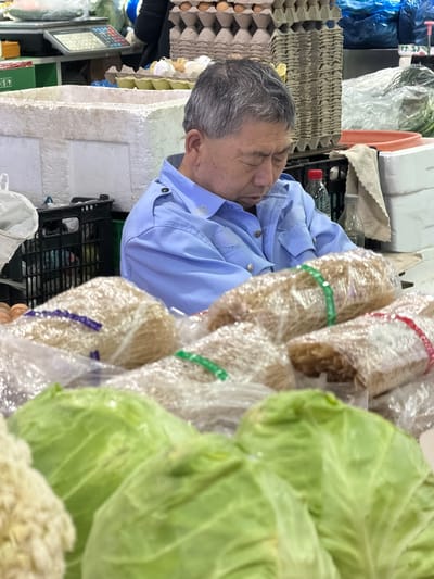 An elderly man with gray hair and a blue shirt is sleeping behind a market stall. The stall contains eggs, cabbages, and packages of dry noodles wrapped in clear plastic. The stall is located in the city of Guta, China.