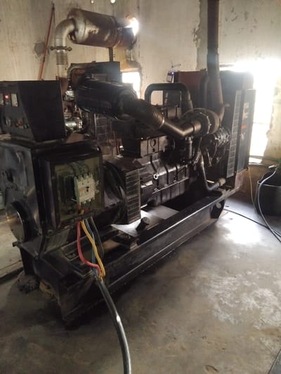 A dark, industrial generator set, likely a diesel engine driven model, sits on a concrete floor in what appears to be a dimly lit indoor space with rough-textured walls. The generator is largely black with various metallic components and visible wiring. A large, cylindrical exhaust pipe, possibly coated in rust, extends upwards and to the left, connected to a flexible joint. In the foreground, a black electrical enclosure is open, revealing a white circuit breaker and multiple colored wires (red, yellow, blue) exiting the enclosure and running down to a thick black cable on the floor. Light streams in from a window or opening to the right, creating a bright, washed-out area and casting reflections on the floor. The general impression is one of a functional, perhaps older, power generation unit in a utilitarian setting. The location in Chhina Uttar, Pakistan, suggests this could be a backup power source for a facility in that region.