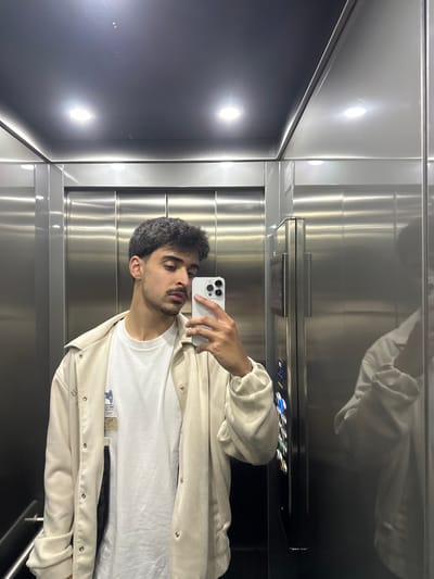 The image displays a young adult male captured in a mirror selfie within an elevator. The subject, positioned centrally, presents dark, short, wavy hair, dark eyebrows, a mustache, and light facial stubble. He wears a white crew-neck t-shirt under an open, light beige or cream-colored long-sleeved jacket secured with snap fasteners. His right hand holds a white smartphone, likely an Apple iPhone with a multi-lens camera array, directed at his reflection.
The elevator interior features brushed stainless steel walls and doors, characterized by a horizontal grain pattern. The dark ceiling contains two brightly illuminated circular recessed lights. A vertical control panel with several illuminated buttons is visible to the right, adjacent to the subject's reflection. The specific location within Guimarães, Portugal, cannot be identified from the visual information provided in the image.
