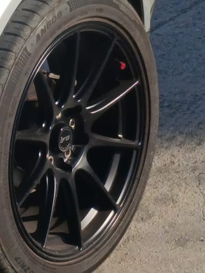 A close-up, diagonal shot captures a black multi-spoke car wheel and tire, likely from a vehicle situated in Polis Chrysochous, Cyprus. The car appears to be stationary, parked on a textured, light-colored paved surface, possibly asphalt or concrete.
The wheel features a sleek black finish with a multi-spoke design and a subtle silver or polished lip. At its hub, a black circular center cap displays a distinctive white or silver "iRa" logo, suggesting the brand of the wheels. Metallic lug nuts are partially visible around the center cap. A notable detail is a small, bright red valve stem cap positioned near the top right of the wheel.
The tire, fitted around the wheel, exhibits clear tread patterns. Several alphanumeric characters are visible on its sidewall: "AN608" is clearly legible, and "ZRIZ" can be seen towards the bottom, likely part of a full tire size or model number (e.g., ZR17). Both the tire and wheel show slight signs of dust or road grime from use. The overall lighting indicates it is daytime.