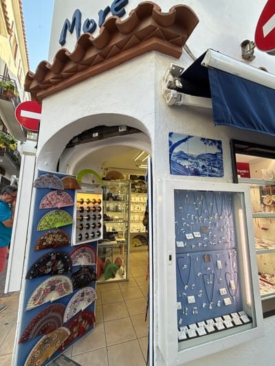 This media file depicts a street scene in Tossa de Mar, Spain, likely during daylight given the bright lighting and clear skies. The focus is on a shopfront displaying various items for sale.
The primary subjects are the merchandise and the shop's exterior. In the foreground, a table is laid out with an assortment of decorative fans, many featuring intricate floral patterns in vibrant colors like pink, green, red, and brown, as well as darker, more subdued designs. Behind the fans, a display case showcases a collection of jewelry, including necklaces, earrings, and pendants, arranged on a blue background. The jewelry appears delicate and possibly made of silver.
The shop itself has a white facade, with a blue awning extending over a section of the entrance. A decorative tiled panel, depicting a blue and white landscape scene, is mounted on the wall. The entrance to the shop leads into a space where more merchandise is visible, including what appears to be additional jewelry displays.
Notable details include a brown, decorative architectural element on the building's exterior, possibly a cornice or a decorative molding. A red and white circular sign is partially visible on the left side of the image. The overall atmosphere suggests a sunny day in a Mediterranean tourist town, with an emphasis on artisanal crafts and souvenirs. There is a glimpse of people in the background, one individual wearing a blue t-shirt. The image is taken from a slightly elevated, angled perspective, looking downwards and across the scene.