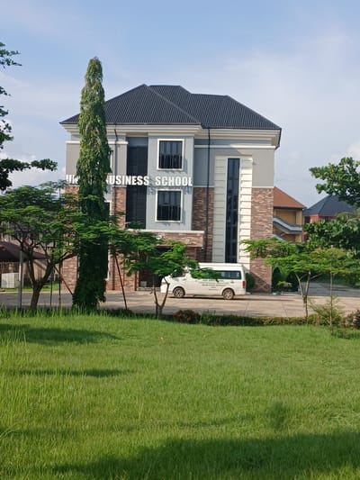 The image shows a modern building with "BUSINESS SCHOOL" written vertically on its facade. The building has a dark, sloped roof and features large windows. It is constructed with a mix of grey panels and brickwork. A white van is parked in front of the building on a paved area. Lush green grass and several trees surround the building, suggesting an outdoor setting. The sky is blue and clear, indicating daytime and fair weather. The scene appears to be in Ukukwa, Nigeria, a location context provided. There is no specific activity or event depicted; it appears to be a general view of the business school. The overall mood is serene and professional. The text on the van is partially visible, but not clearly legible.