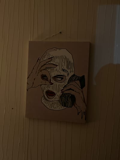 A framed canvas painting depicting a woman wearing a ski mask is hung on a textured, beige wall. The painting shows the woman holding a retro telephone to her ear with her right hand and her left hand is positioned in front of her eye, with her index finger and thumb forming a circle. She has red lipstick and her eyes are visible through the mask. The word "MOTEL" is written on the ski mask near her forehead. The painting is lit by a light source casting a shadow on the right side of the wall. The background is a plain, textured wall, not indicative of any specific urban location.