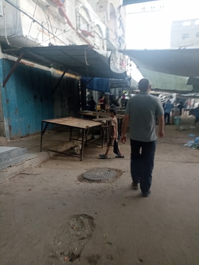 The image depicts a street scene in Gaza City, Palestinian Territories. In the foreground, a man is walking away from the camera, wearing a grey t-shirt and dark trousers. To the left of the man, a young boy in a t-shirt and shorts is walking towards the camera. The environment appears to be an outdoor market or commercial area with stalls and awnings. Various tables and structures are visible, suggesting commercial activity. The ground is paved with concrete, and some debris is present. Buildings line the background, with some appearing to be under construction or renovation. The overall impression is of a busy urban setting.