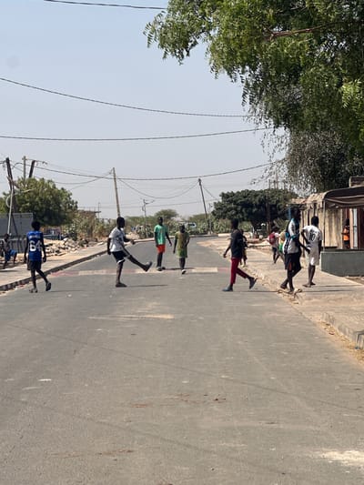 A sunny, daytime shot shows a group of nine children walking/running on a paved road in Communauté rurale de Malicounda, Senegal. The road stretches into the distance, with trees lining the left side. The children are of varying heights and wear different clothing. Utility poles are visible overhead.