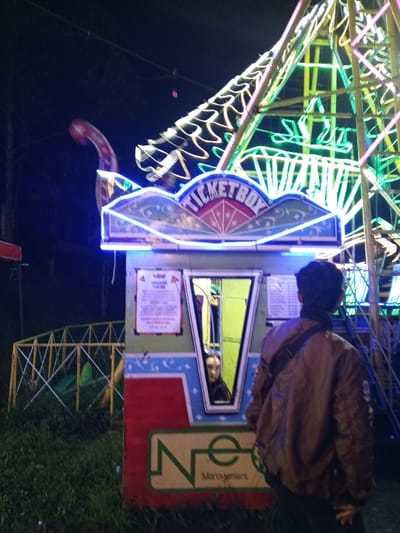 A ticket booth adorned with "TICKET BOX" in blue lights stands beside a brightly lit Ferris wheel at night. The booth is painted with red, blue, and green. A sign is displayed next to the ticket window, and a person is visible inside. A man with a brown jacket and a shoulder bag is standing in front of the booth, his back to the camera. In the background, there is grass and a fence-like structure.