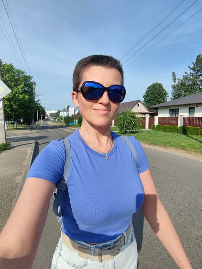 A woman with a short buzz haircut and large, dark sunglasses is captured in a selfie on a paved road in Fanipal, Belarus. She is wearing a vibrant blue t-shirt and light-colored denim shorts, cinched with a beige belt. A light blue backpack is slung over her left shoulder. Her right arm is extended, holding the camera, and her left arm is partially visible. The background features a suburban street with houses, green trees, and utility poles with overhead wires. The sun is bright, casting shadows on the road and the woman's arm. In the reflection of her sunglasses, the selfie-taker is visible along with the surrounding environment.
