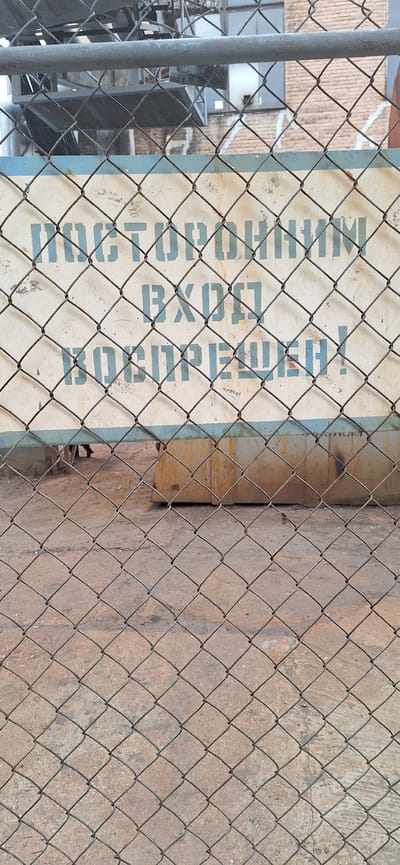 A metal chain-link fence is in the foreground of this image, obscuring a sign and a building. The sign, which is rectangular with a light-colored background and blue text, appears to be mounted on a wooden surface. The text on the sign is partially visible, reading "NATIONAL EXPORT". Behind the sign and fence, a brick building is visible. To the left of the sign, a vertical metal pole, likely part of the fence, is present. To the right of the sign, a textured, reddish-brown surface, possibly a wall or the ground, is visible. The scene appears to be outdoors, with natural lighting. There are no people visible in the image. The context provided indicates the location is Nakhodka, Russia.