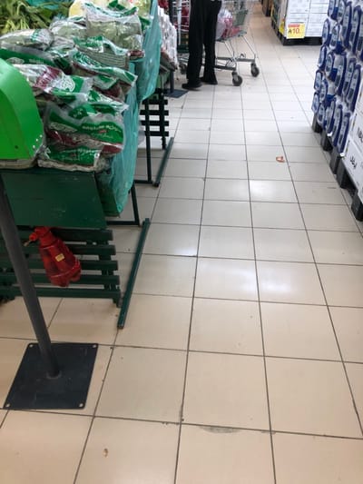 The image displays an interior view of a supermarket aisle located in Riyadh, Saudi Arabia. The flooring consists of light-colored square ceramic tiles, showing signs of wear, including scuffs, dirt accumulation, and minor chips. Artificial overhead lighting illuminates the scene.
On the left side of the aisle, a green metal display stand, partially draped with a light green plastic sheet, is stacked with multiple clear plastic bags containing leafy green produce, identified by visible Arabic text as "Molokhia". Beneath this stand, a red plastic spray bottle rests on a lower shelf. A black metal pole with a square base is positioned in the foreground on the left.
On the right side of the aisle, an array of packaged goods, likely cartons, featuring blue and white branding, is neatly stacked on black pallets or shelving units. A yellow price tag displaying the number "10" is visible on a higher shelf.
In the mid-ground, the lower legs and feet of one individual are visible, clad in dark pants and dark shoes, standing behind a silver metal shopping cart. A small, light-brown piece of debris is present on the floor tiles in the center of the aisle. No direct interaction or specific action by the individual is discernible beyond their presence with the shopping cart.