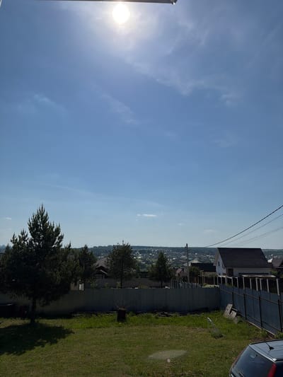 The photograph depicts a residential area on a sunny day. A house with a dark roof is visible, alongside a metal fence. Green grass covers the ground adjacent to the house. In the background, a cityscape stretches into the distance under a blue sky with scattered clouds. Utility poles and wires are visible above the houses. Based on location data, this scene is in Ягул, Russia. A blue car is parked to the right of the frame.