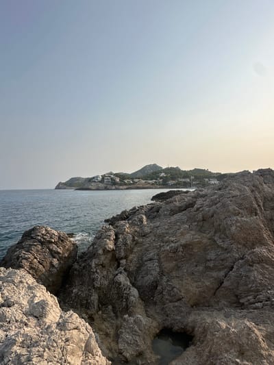 The image presents a panoramic coastal scene located in Font De Sa Cala, Spain. The foreground is dominated by rugged, grey-brown rock formations, characterized by sharp angles, visible fissures, and an uneven texture. A small, irregular depression in the rocks in the lower right contains a pool of still water. The midground features calm, dark blue-green sea water extending from the rocky shore towards the horizon. In the background, a distant headland extends into the sea on the left. To the right, a verdant hillside rises from the coast, dotted with numerous white-walled buildings, indicative of a populated area. Behind these structures, a range of prominent, rocky mountains is visible, with a faint, slender tower-like object positioned on one of the distant peaks. The sky above is light blue on the left, transitioning to a hazy pale yellow-white on the right. No people, actions, or direct interactions are depicted within the frame.