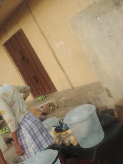 The image depicts an outdoor or semi-outdoor scene in Neni, Nigeria, against a light yellow-ochre painted building wall with signs of wear and a closed, corrugated brown metal door. A concrete or stone platform runs along the base of the wall.
In the mid-ground, one individual is partially visible from the waist up, facing away from the camera and leaning forward. They are wearing a light-colored, long-sleeved shirt patterned with yellow lemons and white ducks, over a blue and white checkered skirt or wrapper.
On the ground and platform in the foreground, various items are present:
* Several green, ovoid fruits or vegetables are scattered.
* A silver-colored basin is partially visible on the ground near the individual.
* Two transparent plastic buckets with lids are in the foreground; one in the lower-left, partially obscured, and another more prominent in the lower-right, containing clear liquid.
* Behind these buckets, on a dark surface, are multiple clear plastic bags containing pale yellow or white solid items, along with several clear plastic bottles with yellow caps.
* A pair of dark-colored footwear is also on the dark surface.
* A black plastic bag is positioned near the rightmost bucket.
* A light-colored bag is on the platform near the corrugated door.
The arrangement of items suggests an activity involving food preparation, sorting, or an informal vending setup.