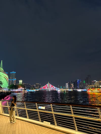 A night scene in the Yuexiu District of Guangzhou, China shows a waterfront view. The Pearl River reflects colorful lights from buildings, including a bridge adorned with red floral patterns and a partially-visible green domed structure. A woman stands on a wooden boardwalk behind a light-colored railing, looking at the water. The sky is dark and cloudy.