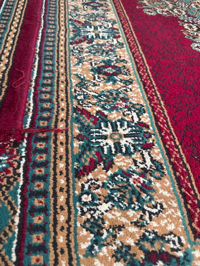 The image is a close-up, angled shot of a patterned rug. The rug features a repeating geometric design with stripes and floral motifs in colors of maroon, teal, black, white, and tan. A thin, red thread is visible on the left side of the rug, appearing to be a loose end or a frayed section. The background is out of focus, emphasizing the texture and pattern of the rug. The location is not specified in the image or the prompt.
