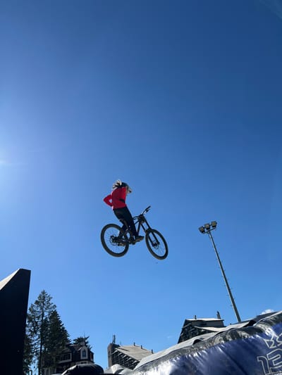 A person is captured mid-air on a mountain bike, performing a jump against a clear blue sky. The cyclist, wearing a red jacket and helmet, is positioned centrally in the frame. The bicycle appears to be a downhill mountain bike, with knobby tires and suspension. In the background, several buildings are visible, including what appears to be a ski resort or a mountain lodge, suggesting an outdoor recreational area. There is also a tall pole with lights, likely for evening events or lighting up a ski slope. The image seems to be taken at a location associated with outdoor sports or events, possibly in Samokov, Bulgaria, given the context of a ski resort. The perspective is from below, looking up at the cyclist and the sky.