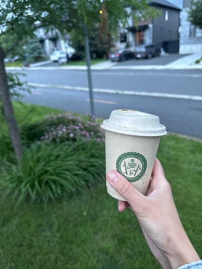 A close-up shot shows a hand holding a disposable coffee cup made of a light brown, biodegradable material. The cup has a white lid and features a green circular logo with text and an illustration of bamboo stalks. The background is blurred, but it appears to be an outdoor setting with a street, cars, and houses visible. The overall impression is a casual moment, possibly in a residential area of Gatineau, Canada, given the architectural style of the houses and the presence of mature trees and manicured lawns. The lighting suggests it is either early morning or late afternoon.