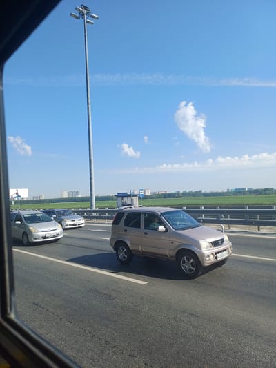 An eye-level, outdoor, daytime shot captures a multi-lane highway with a significant traffic flow under a bright blue sky. The lane nearest the foreground has two silver cars: a hatchback on the left and a sedan on the right. Further ahead in the same lane, a beige SUV is driving. In the adjacent lane, a second silver sedan is visible. The highway is lined with a metal guardrail separating it from a verdant, flat field. In the background, a row of residential buildings and some industrial structures are visible. Dominating the sky is a very tall, multi-headed light pole, characteristic of stadium or highway lighting. A few wispy clouds are scattered across the sky. The image appears to be taken from a vehicle, given the slight framing and blur on the left side, suggesting a window view. The location is identified as Elitnyi, Russia.