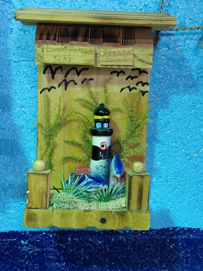 A rustic, wooden wall decoration, likely found in Cutler Bay, United States of America, depicts a stylized lighthouse scene. The piece is constructed from various wooden scraps, arranged to form a small diorama. A ceramic lighthouse model, painted with black, white, red, and blue sections, lies horizontally within a central opening, suggesting it might represent a fallen or capsized lighthouse. Patches of artificial moss and green, sprig-like foliage are scattered around the lighthouse, possibly intended to represent land or vegetation. Black painted "V" shapes, possibly meant to represent seagulls, are visible on the wooden backdrop. To the left of the lighthouse scene, a small wooden block is labeled "Lighthouse Bar Frozen Drink", indicating a theme of a seaside bar or establishment. The entire assemblage is mounted on a rectangular wooden base with a hanging rope at the top. The backdrop against which the piece is displayed is a blue, textured surface, with a darker blue section visible on the right.