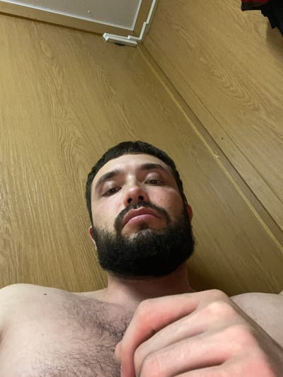 An eye-level, low-angle, close-up shot shows a shirtless man with a beard, viewed from the chest up. The man is looking directly at the camera, with his face tilted slightly upwards. His skin is fair, and he has dark hair. The background consists of a wooden wall with a horizontal grain pattern. There are also some white rectangular objects mounted on the wall near the top of the frame. The lighting appears to be artificial, casting shadows on the man's face and chest.