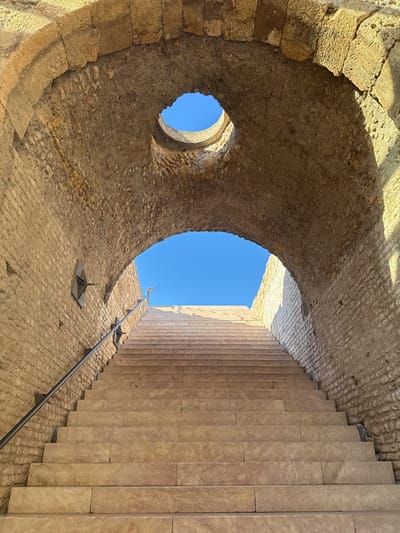 A low-angle shot showcases stone stairs leading upwards through a weathered tunnel-like structure in Tarragona, Spain. The walls of the tunnel, constructed from rough-hewn stone, converge in an arched ceiling. A circular opening, a sort of oculus, is visible in the ceiling. Sunlight illuminates the steps and walls, creating sharp contrasts. A metal handrail runs along the left side of the stairs. The blue sky is visible through the openings at the top of the stairs and in the circular opening above.
