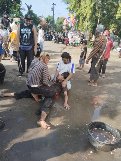 This outdoor image, taken in Sragen, Indonesia, captures a central activity surrounded by a gathering of people under bright daylight. In the foreground, a person is lying on a muddy, wet ground, partially submerged in a puddle. Two men are attending to this individual: one, with a striped shirt and a headband, kneels on the lower body, while another, wearing a white and blue polo shirt, cradles the head and upper torso. A black basin filled with water and pink and white flower petals sits nearby in the puddle, suggesting a ritualistic bathing or cleansing.
Around this central scene, several bystanders observe. Notable figures include a man in a black sleeveless shirt and cap, wearing a blue fanny pack, whose shirt features text resembling a band logo (partially visible text like "WALKGEN OF CITY"). In the background, a street vendor sells colorful balloons and other items from a cart, while other people and motorcycles are visible. The ground is a mix of dirt, mud, and water. The overall atmosphere is one of observation at what appears to be a public performance or cultural ritual, rather than an emergency. The clear sky and strong shadows indicate it is mid-morning or afternoon.